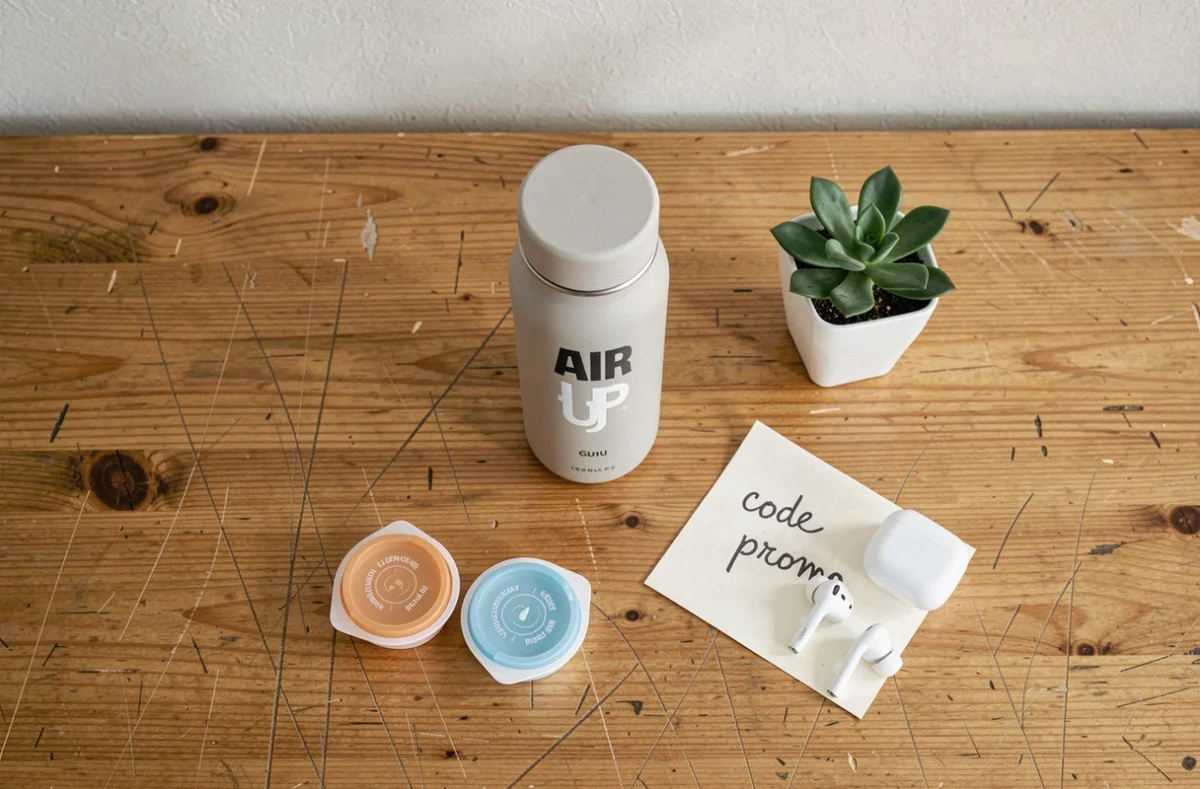 Flat-lay d'une bouteille Air Up avec pods aromatiques, note manuscrite avec code promo et accessoires sur un bureau en bois